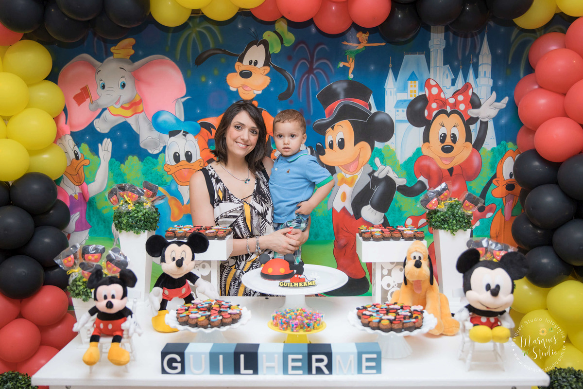 Foto tirada em um buffet localizado na Pompeia - São Paulo, SP no aniversário de dois anos do Guilherme. Na foto temos o aniversariante no colo da sua mamãe, eles estão fazendo pose e se encontram atrás da mesa do bolo. 
