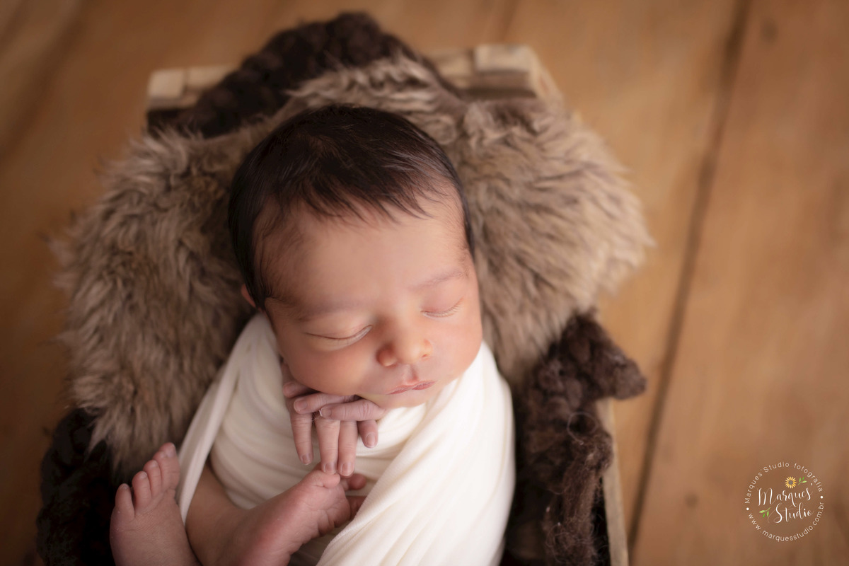 Fotografia registrada no ensaio Newborn do Bernador . Na foto o bebê está deitado em um  caixote de madeira, em cima de um tecido marrom. Ele está enrolado em um wrap bege. O fundo fotográfico é de madeira. Sutido localizado em Perdizes - São Paulo, SP