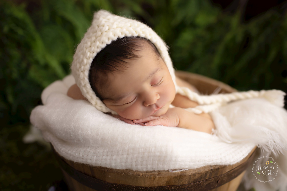 Fotografia registrada no ensaio newborn do Bernardo. Na foto temos o bebê deitado em um balde de madeira, coberto com uma manta branca e um gorro branco na cabeça. O fundo fotográfico é de um lindo e verde jardim. Studio está na Zona Sul de São Paulo, SP.