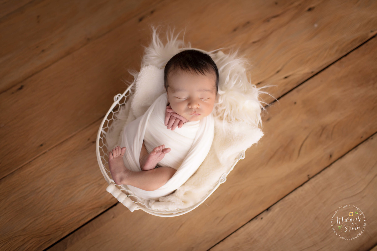 Foto registrada em studio localizado na Av. Paulista - São Paulo, SP. Na foto temos um récem nascido menino, deitado em uma cestinha branca, coberta por um tecido fofo branco. Ele esta enrolado em wrap de cor bege, de barriga pra cima, 