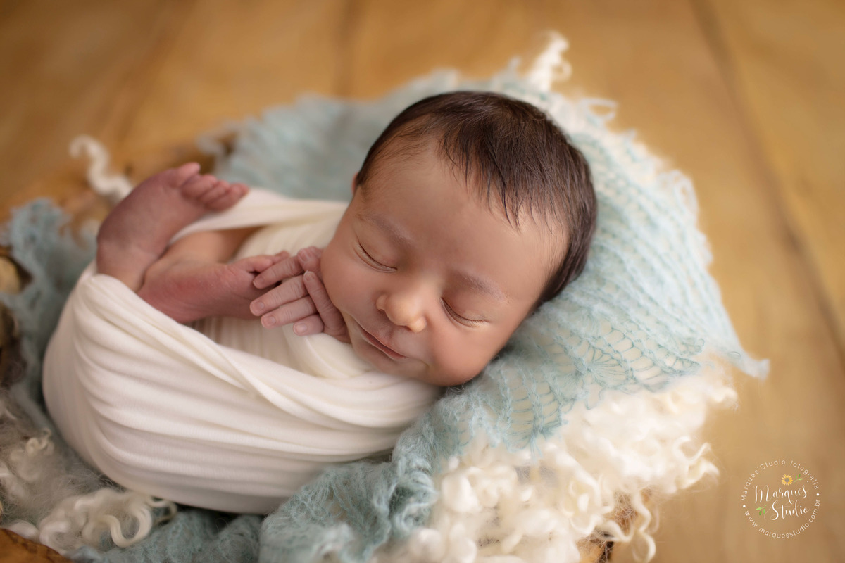 Fotografia tirada em studio localizado na Pompeia - São Paulo, SP. Na foto temos um récem nascido menino, deitado em uma cesta de madeira, coberta por um tecido branco e outro azul. Ele esta enrolado em wrap de cor bege, deitado de ladinho.