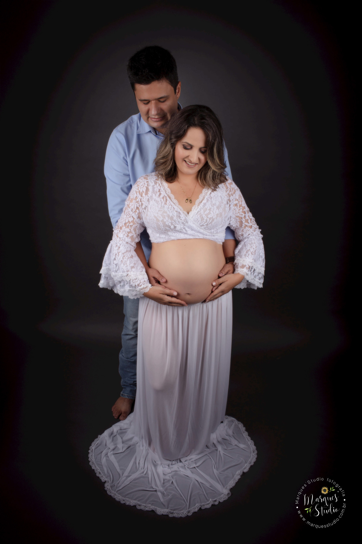 Ensaio de gestante da Sara realizado no studio que fica localizado na Av. Consolação - São Paulo, SP. Na foto temos um casal de grávidos se abraçando e segurando a barriga da mamãe. Ela está com um conjunto branco de saia e blusa, e ele de camisa social. 