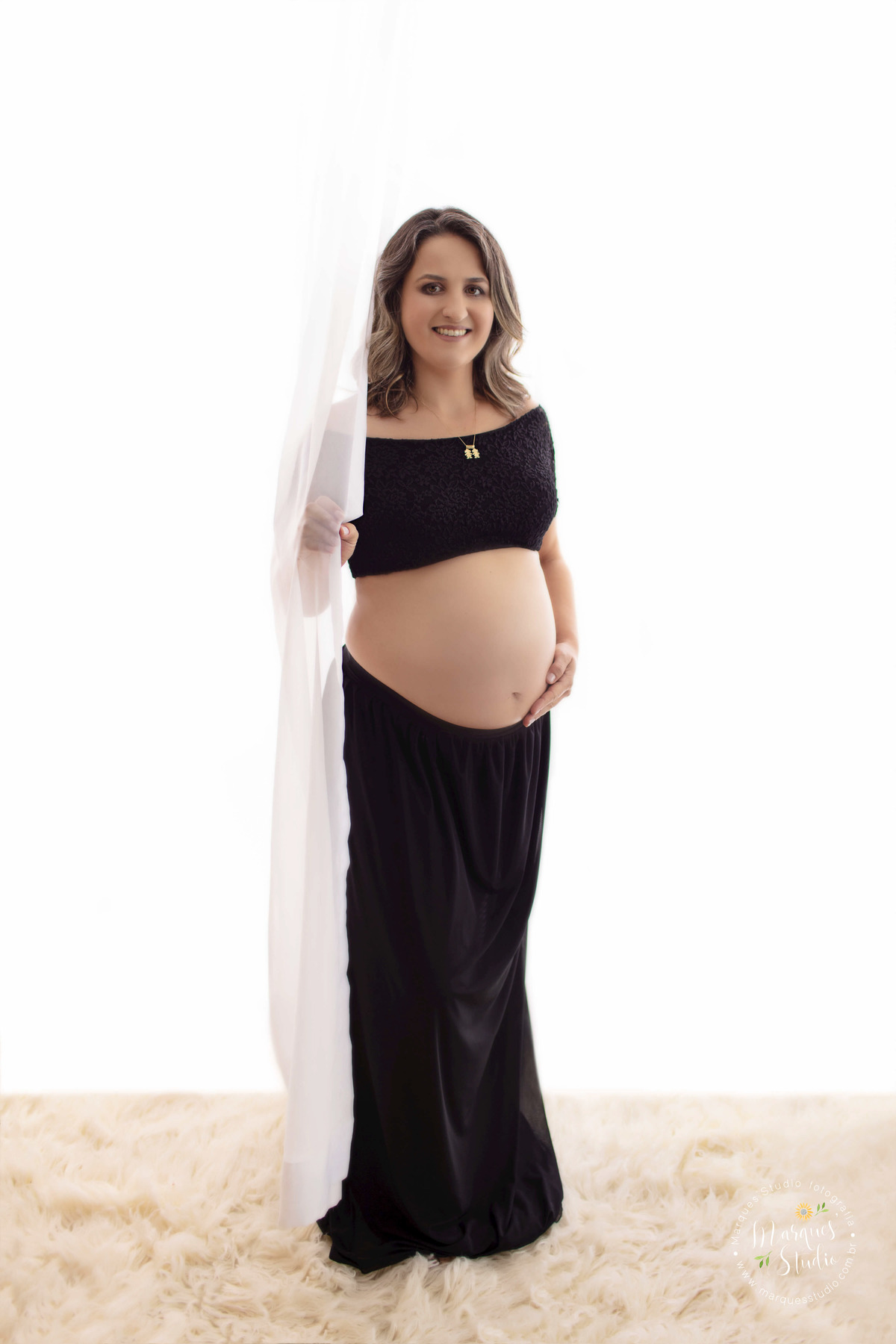 Fotografia registrada no ensaio fotográfico da Sara no studio que fica localizado em Santo Amaro - São Paulo, SP. Na foto temos a mamãe usando um conjunto preto de saia e blusa, segurando com a mão direita uma cortina branca e com a esquerda a barriga.