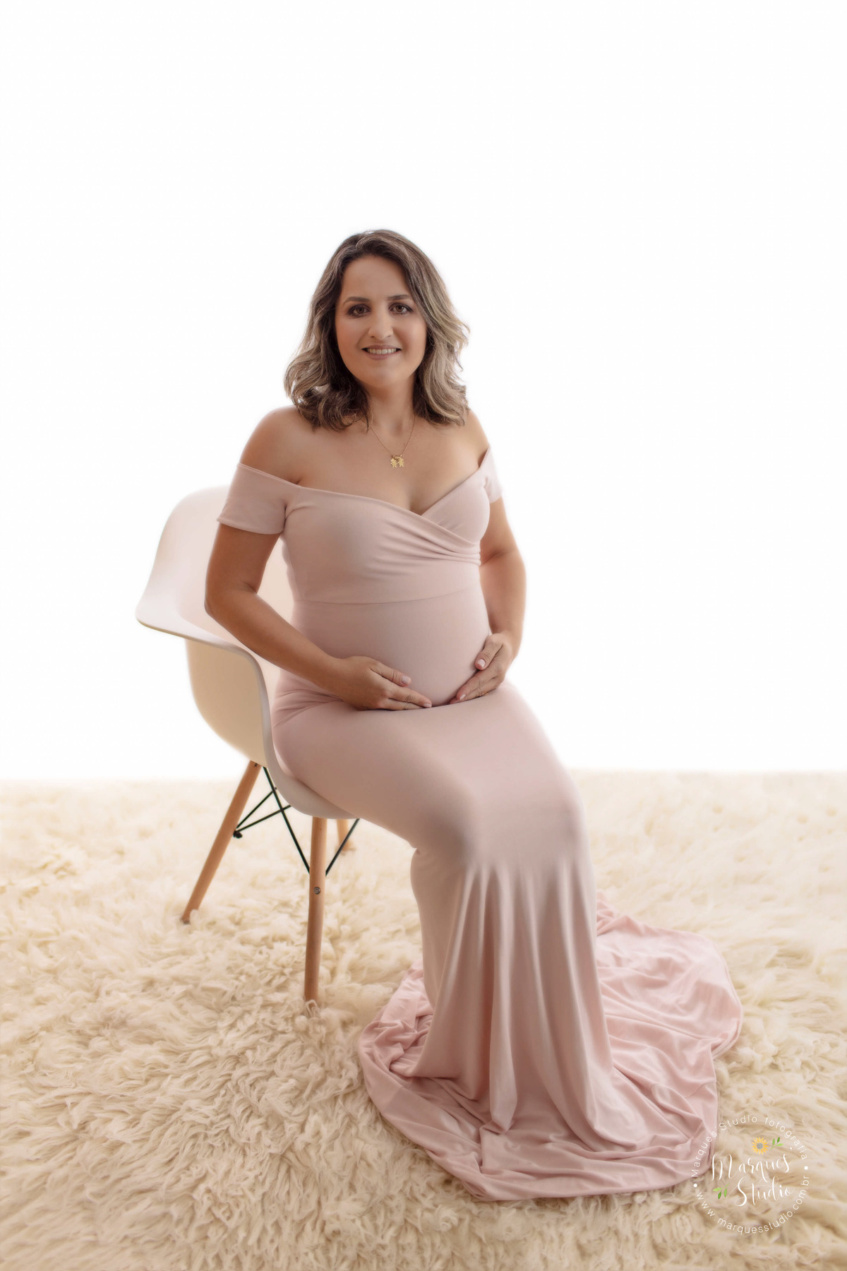 Ensaio de gestante feito em studio localizado na Vila Leopoldina - São Paulo, SP. Na foto temos a mamãe Sara sentada em uma cadeira branca, em cima de um tapete bege, vestindo um lindo vestido rosê com as mãos na barriga e sorrindo.