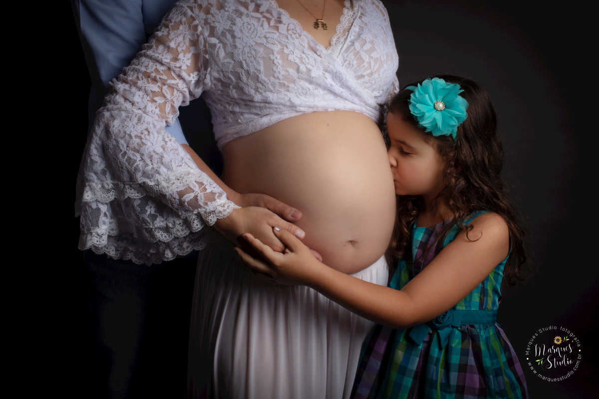 Ensaio de gestante feito em studio que fica localizado na Zona Sul de São Paulo, SP. Na foto temos um close da barriga da mamãe sendo abraçada pelo papai e a sua filinha mais velha dando um beijo carinhoso nela. 