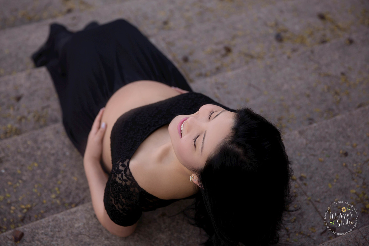 Foto feita no ensaio fotográfico da Karla. Na foto a mamãe está sentada em uma escada, vestida com um lindo conjunto preto de saia e blusa, com as mãos na barriga, sorrindo. O ângulo está de cima para baixo. Foto tirada na Vila Leopoldina - São Paulo, SP.