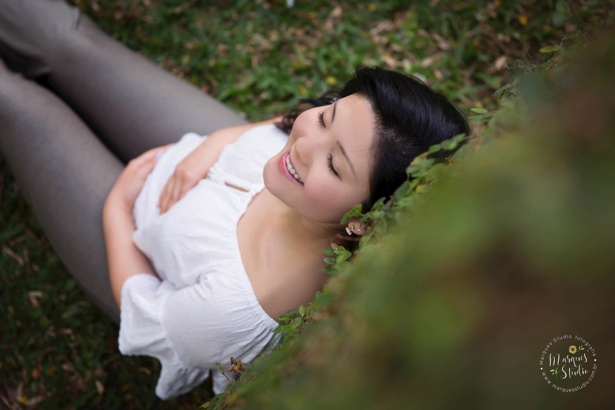 Foto feita no ensaio fotográfico da Karla. Na foto a mamãe está sentada em um jardim, vestida com uma linda blusa branca, com as mãos na barriga e sorrindo serena. O ângulo está de cima para baixo. Foto tirada na Vila Leopoldina - São Paulo, SP.