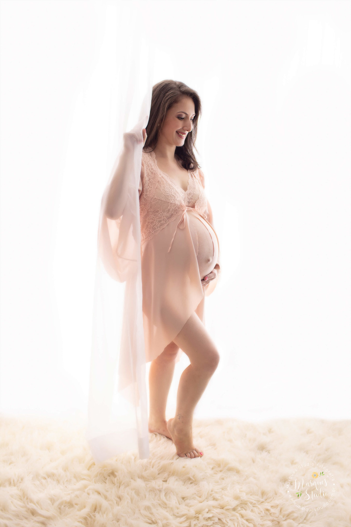 Ensaio de gestante da Eliene em studio localizado na Av. Consolação - São Paulo, SP. Na foto temos a futura mamãe atrás de um cortina branca, vestida com uma linda camisola rosê. Sua mão direita segurando a cortina e a esquerda sua barriga de gestante.