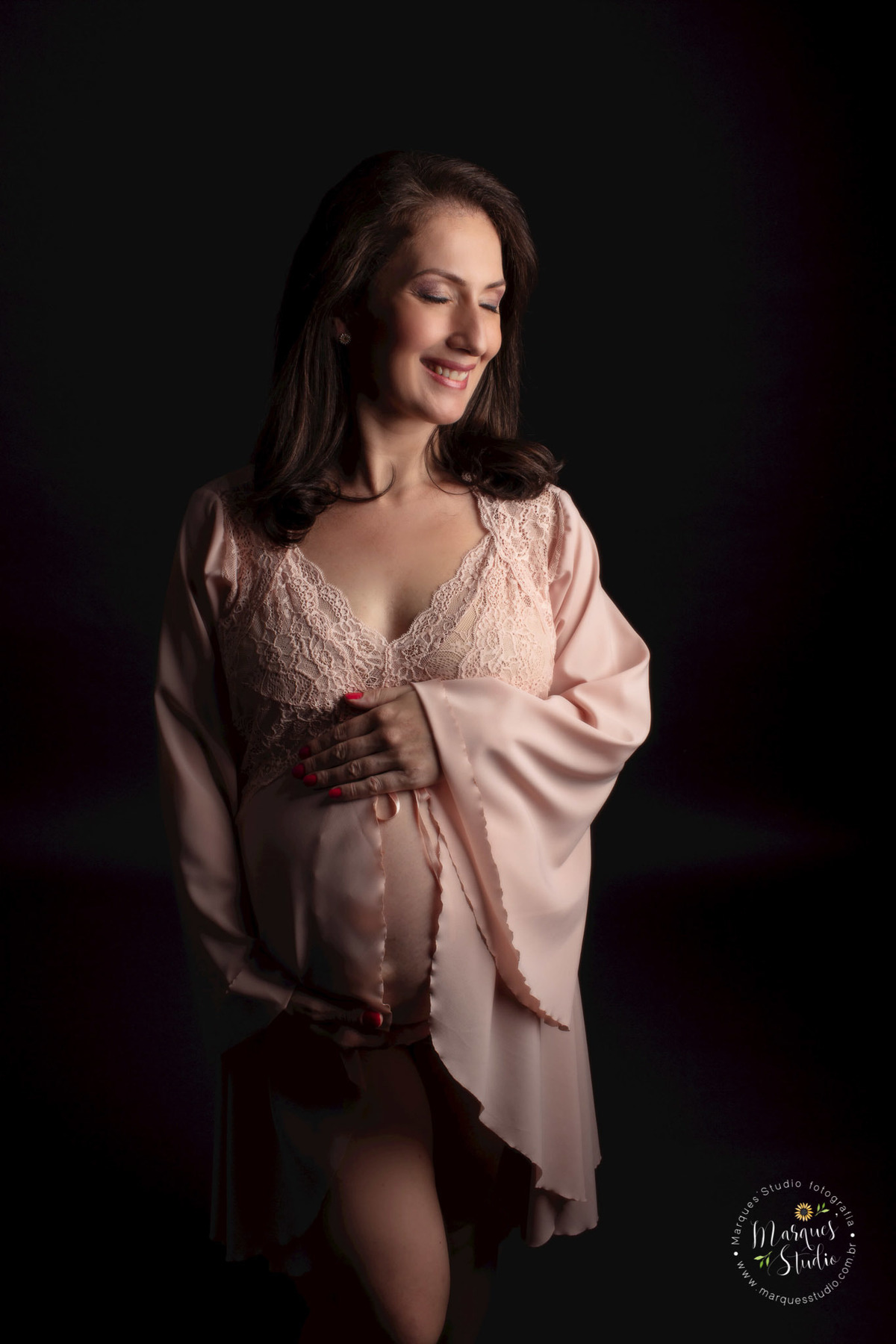 Ensaio de gestante da Eliene em studio localizado em Perdizes - São Paulo, SP. Na foto temos a futura mamãe vestida com uma linda camisola rosê, olhando para o lado e sorrindo, com as suas mãos na sua barriga de gestante.