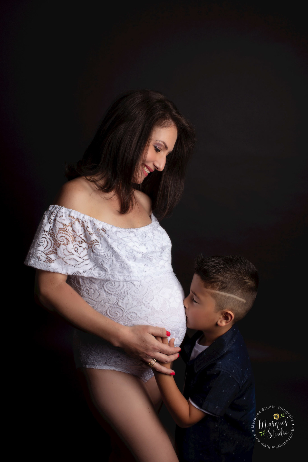 Ensaio de gestante da Eliene em studio localizado na Vila Madalena - São Paulo, SP. Na foto o filhinho mais velho está dando um beijo na barriga da mamãe. Ele está vestindo um lindo bory branco, olhando pra ele e sorrindo. 