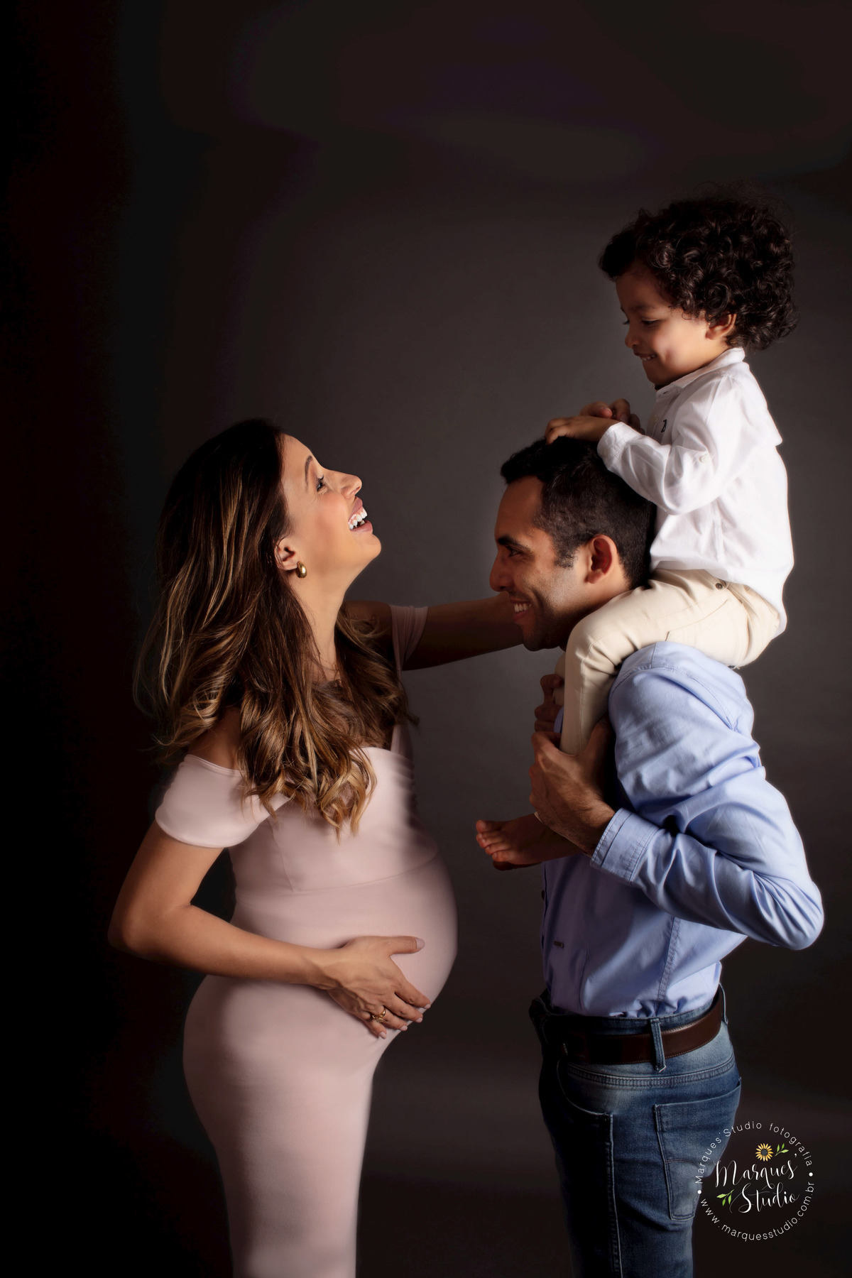 Ensaio de gestante da Nira de 32 semanas. Na foto temos o seu filho mais velho sentado nas costas do papai que está sorrindo, enquanto a mamãe brinca com ele e sorri. O studio onde foi feita a foto encontra-se na Zona Oeste de São Paulo, SP