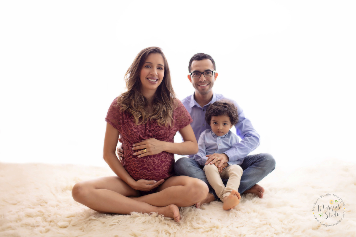 Fotografia tirada no ensaio de gestante da Nira com 32 semanas. Na foto temos toda a família sentada em um carpete bege. O papai com o filhinho no colo e a mamãe com as mãos na barriga. O studio fica localizado em Perdizes - São Paulo, SP.