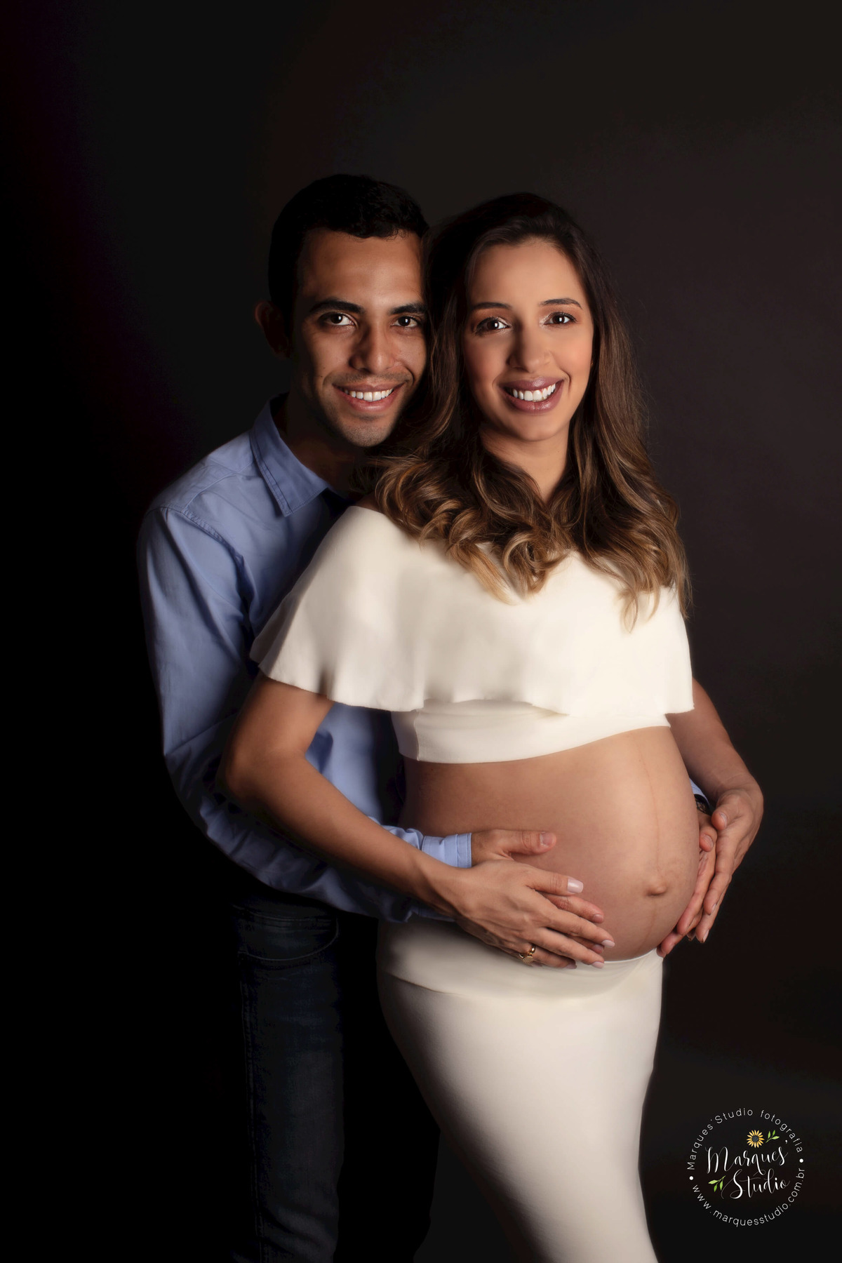 Foto registrada no ensaio de gestante da Nira. Na foto temos ela e o marido em um abraço segurando a barriga, Eles estão sorridentes e felizes. Ela está usando um conjuntinho bege e ele camisa azul social. Studio encontra-se na Zona Oeste de São Paulo, SP