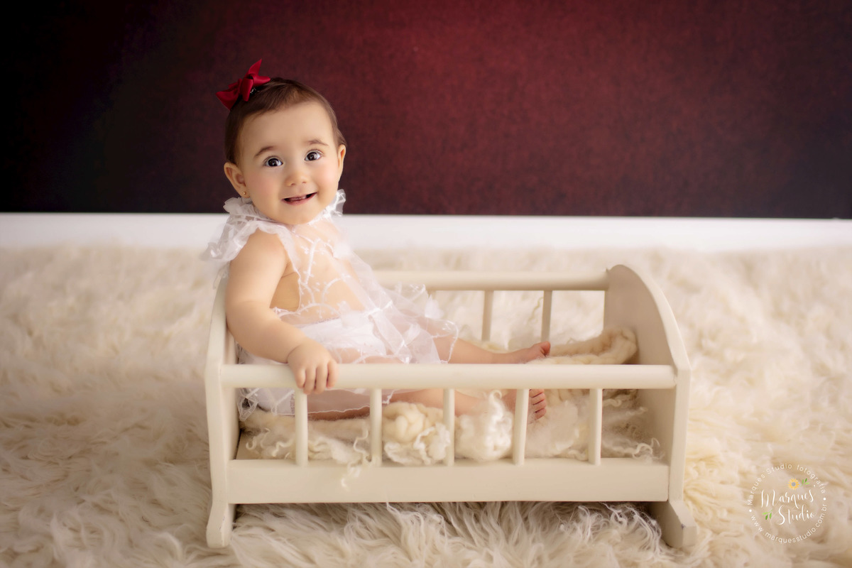 Ensaio feito em studio que fica localizado na Vila Madalena - São Paulo, SP. Na foto temos a Bia, uma bebê de 9 meses, senta da um berço bege, usando um vestido branco transparente e um lacinho vermelho na cabeça. Ela está sorrindo. 