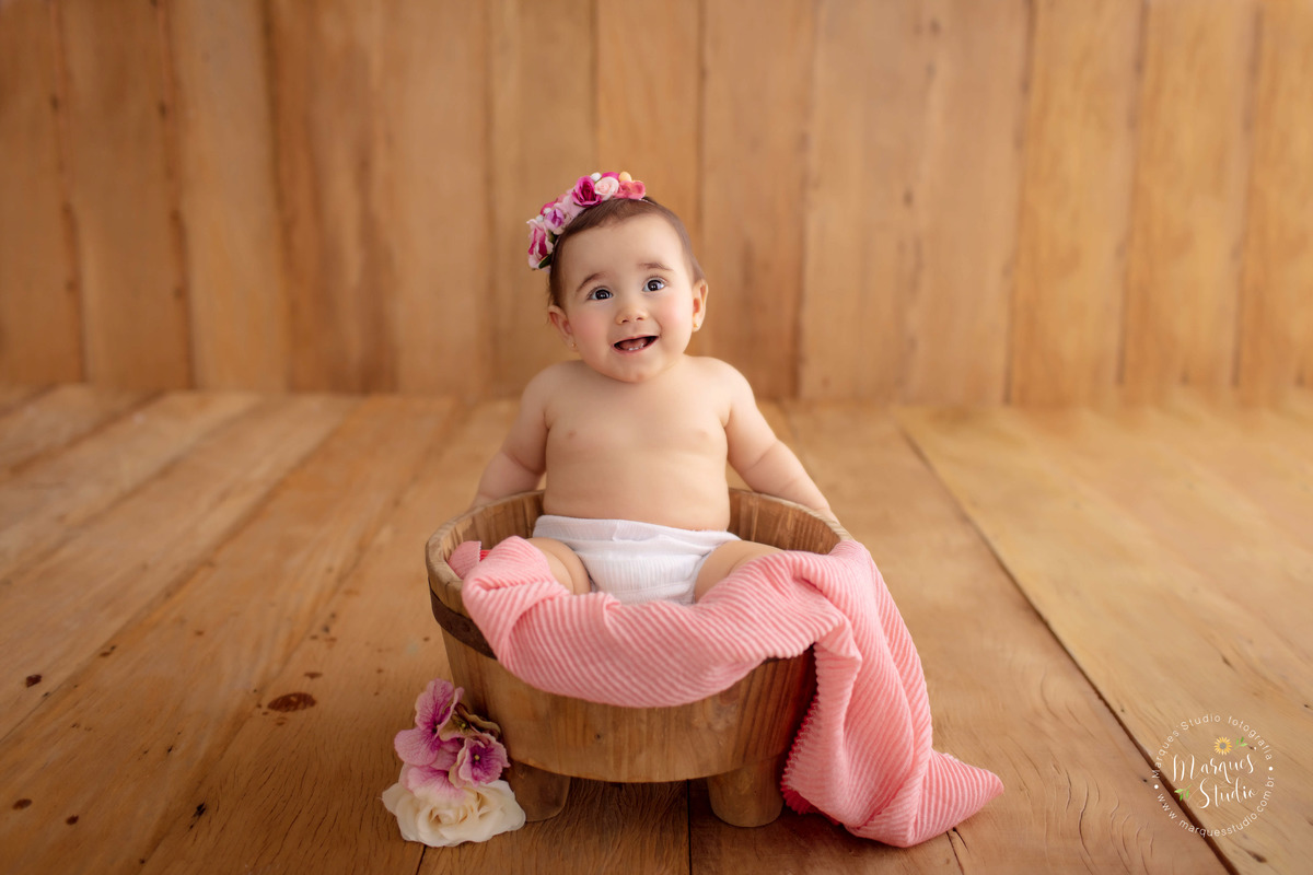 Ensaio de bebê da Bia de 9 meses. Na foto ela está sentada em um balde de madeira, com uma manta rosa e flores em volta. Ela usa flores na cabeça e está com um sorriso fofo no rosto. O studio fica localizado em Santo Amaro - São Paulo, SP. 