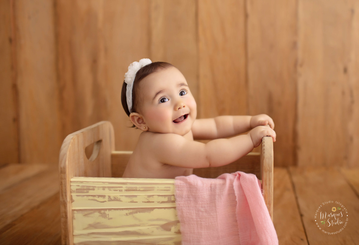 Ensaio de bebê da Bia de 9 meses. Na foto ela está sentada em um berço de madeira, com uma manta rosa. Ela usa uma faixa branca na cabeça e está com um sorriso fofo no rosto. O studio fica localizado na Pompeia - São Paulo, SP. 