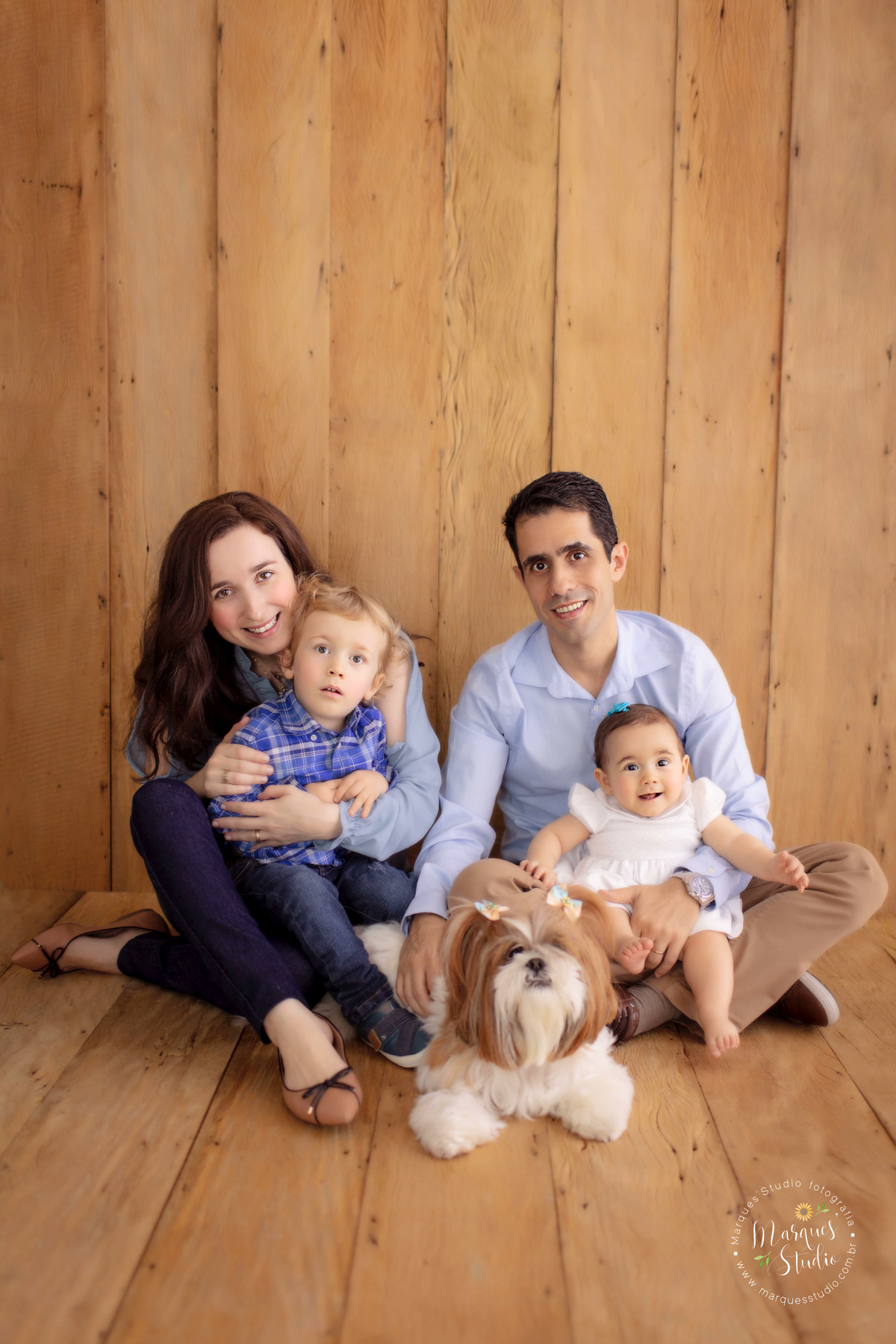 Ensaio feito em studio que encontra-se localizado na Av. Paulista - São Paulo, SP. Na foto temos a mamãe sentada no chão com o filho mais velho no colo, o papai sentando no chão com a bebê no colo e a cachorra da família no meio, todos sorriem felizes.