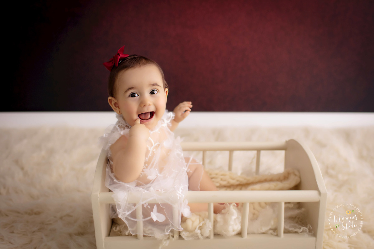 Foto registrada em studio que fica localizado em Perdizes - São Paulo, SP. Na foto temos a Bia, uma bebê de 9 meses, senta da um berço bege, usando um vestido branco transparente e um lacinho vermelho na cabeça. Ela está sorrindo com o dedo na boca.