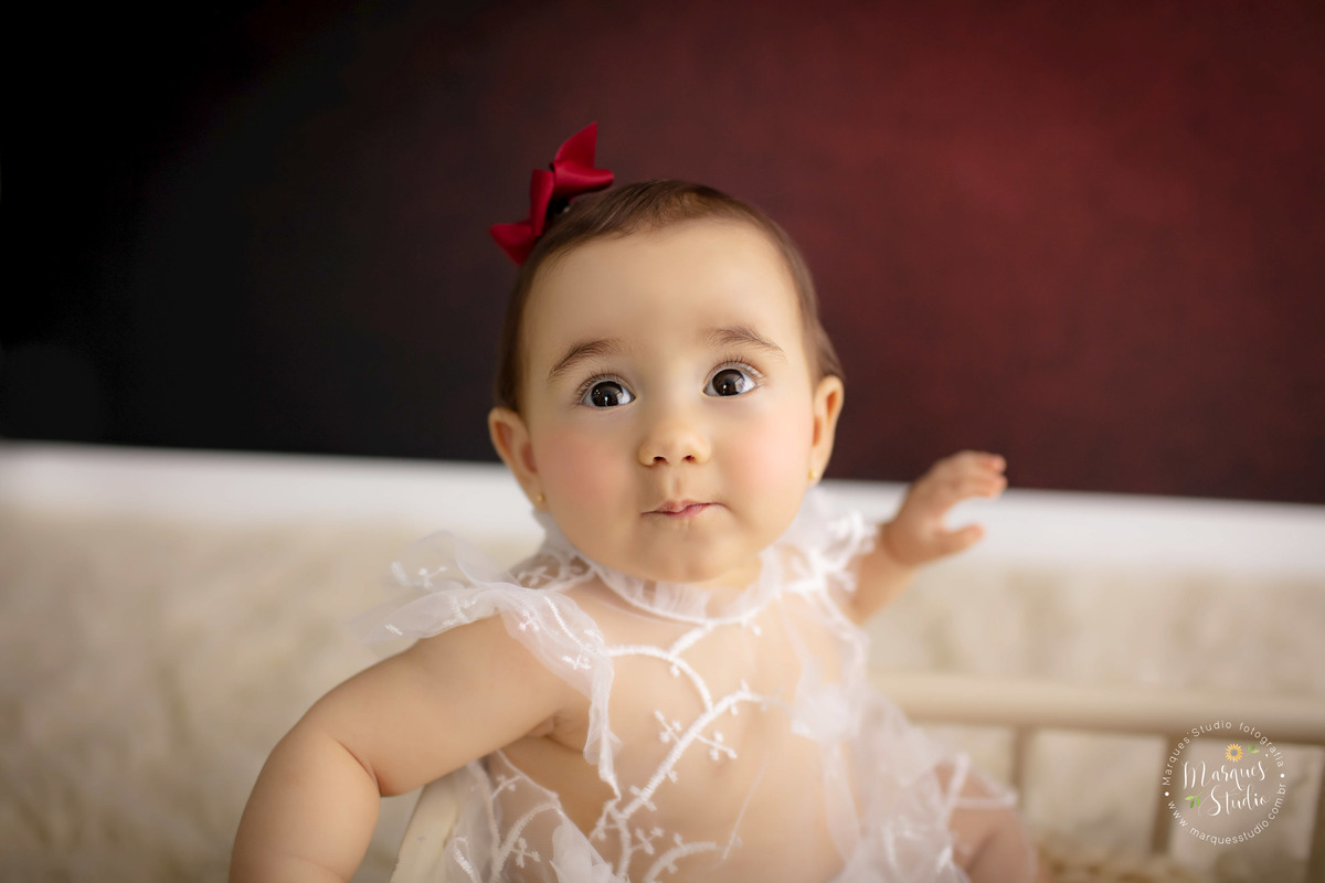 Foto feita no ensaio de 9 meses da Bia. Na foto temos um lindo close no rosto da bebê, que está fazendo biquinho, ela usa um vestido branco transparente e um laço vermelho na cabeça. Studio fica localizado na Vila Leopoldina - São Paulo, SP.