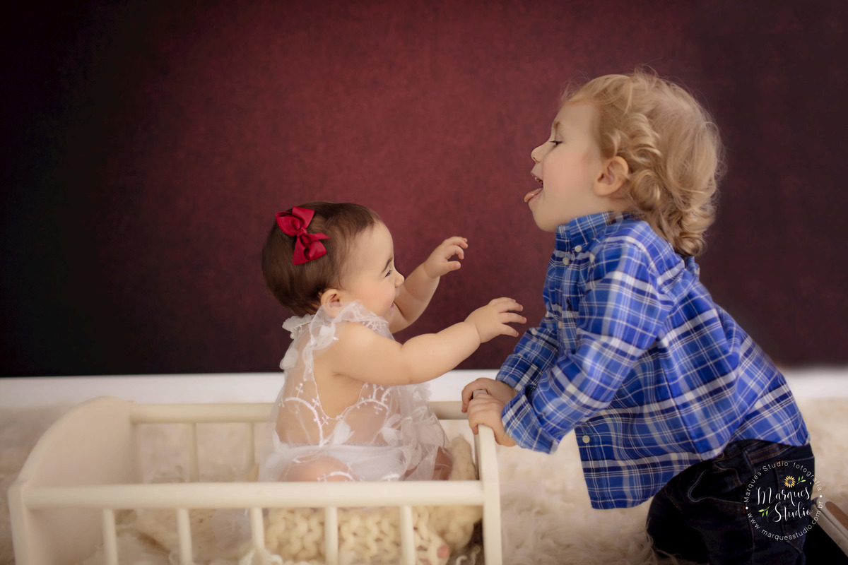 Fotografia registrada no ensaio de 9 meses da Bia. Na foto temos a Bia no berço bege e o seu irmão mais velho brincando com ela de fora. Eles estão rindo e brincando. Studio localizado na Av. Paulista - São Paulo - SP.