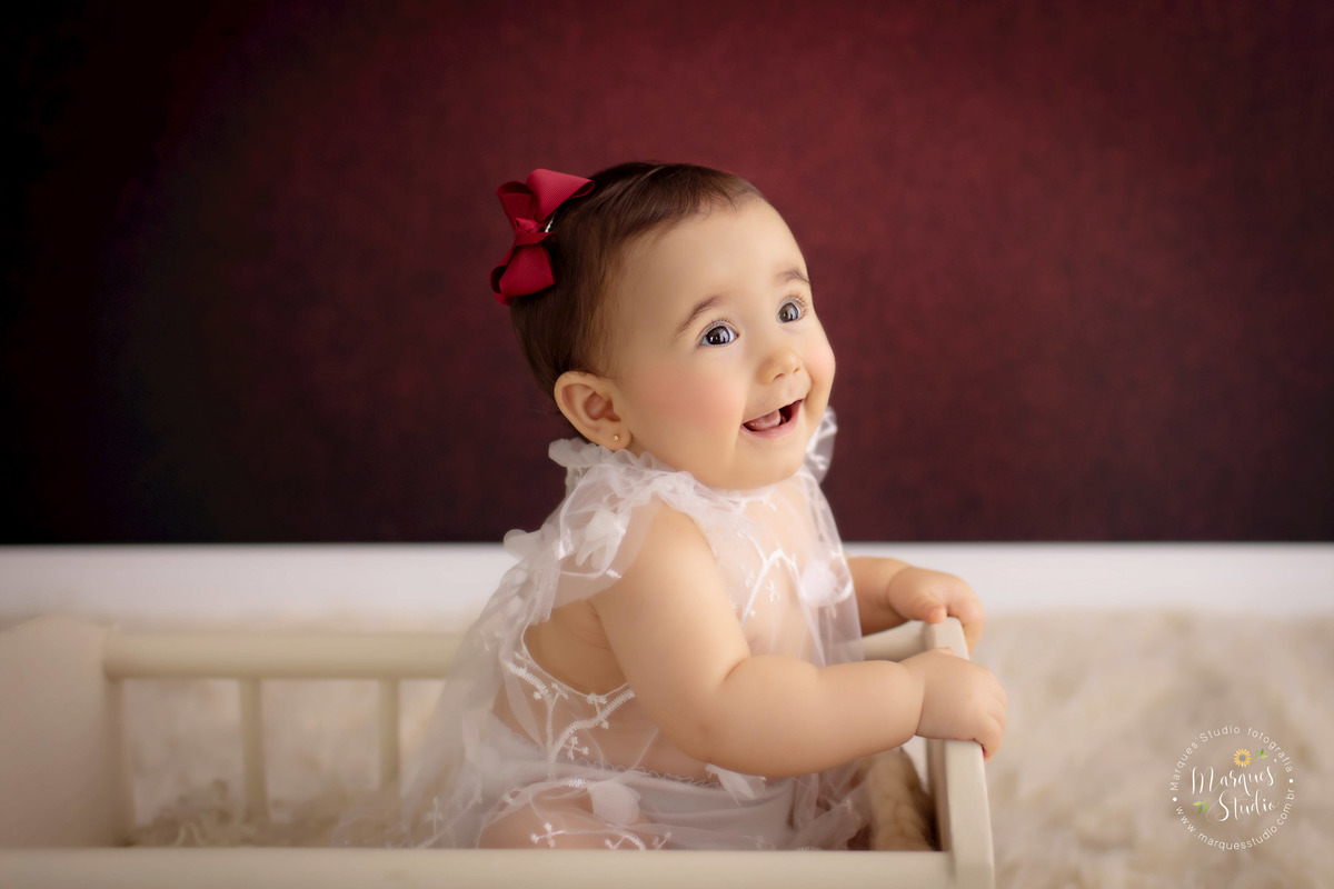 Ensaio feito em studio que fica localizado na Av. Consolação - São Paulo, SP. Na foto temos a Bia, uma bebê de 9 meses, senta da um berço bege, usando um vestido branco transparente e um lacinho vermelho na cabeça. Ela está com um lindo sorriso no lábio. 