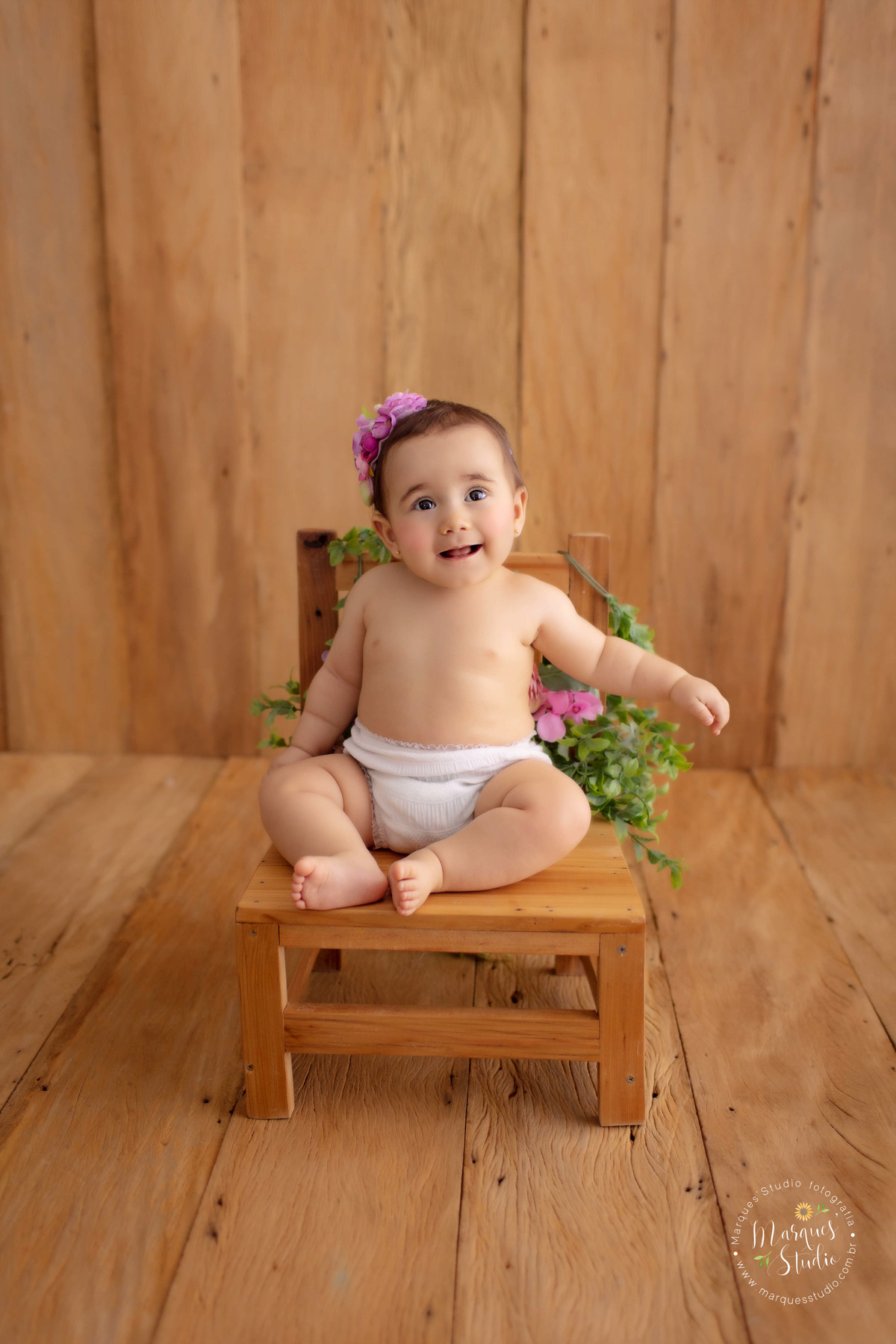 Foto feita em studio que se encontra localizado em Osasco - São Paulo, SP. Na foto temos a bebê Bia, com um flores roxas na cabeça, usando uma calcinha branca e sentada em uma cadeira de madeira que contém um mini jardim. Ela está sorrindo de forma fofa.