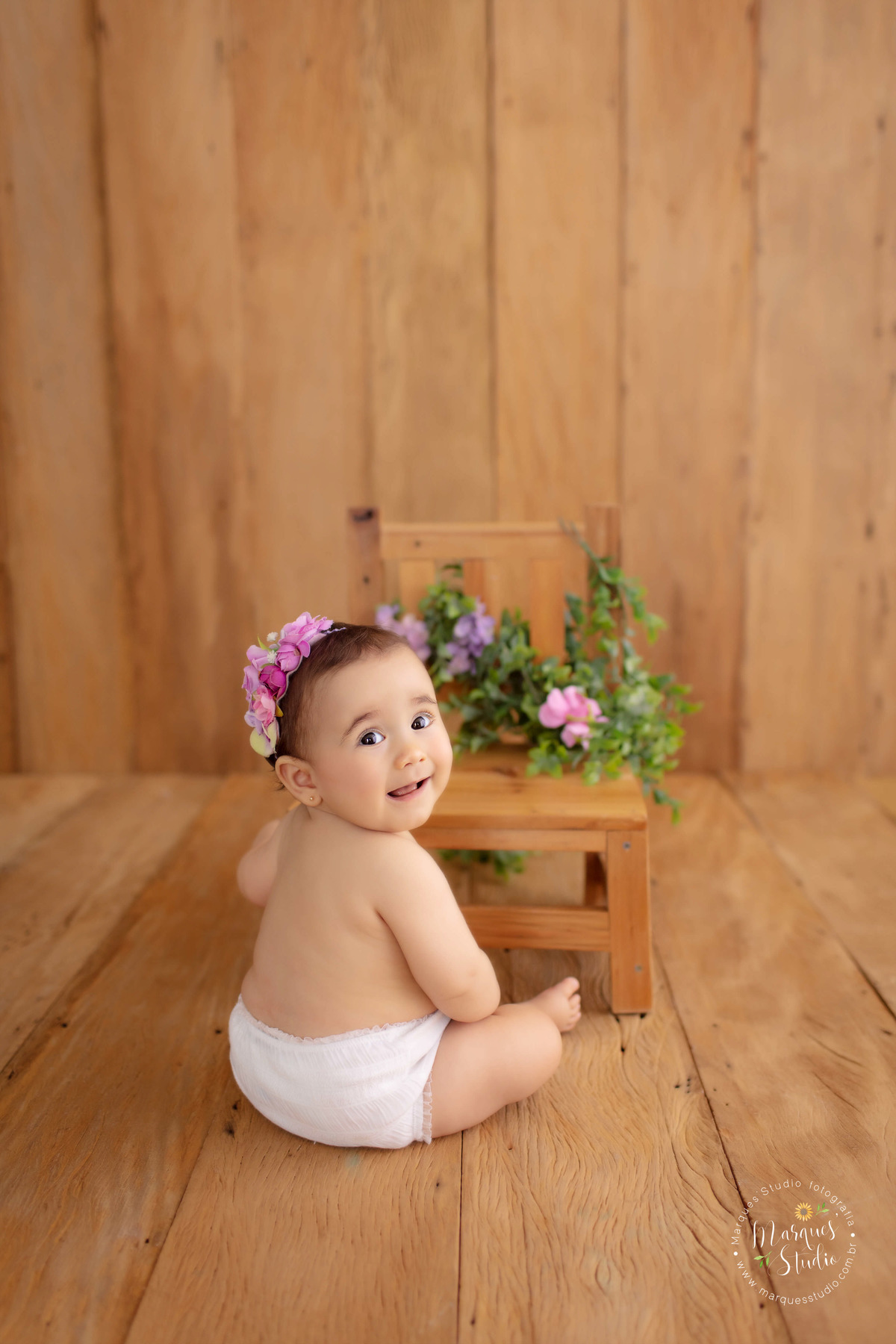Fotografia realizada no ensaio de 9 meses da Bia em studio que encontra-se localizado na Zona Oeste de São Paulo, SP. Na foto a Bia está sentada no chão, de costas e olhando pra câmera sorrindo. No cenário temos uma cadeira de madeira com um mini jardim. 