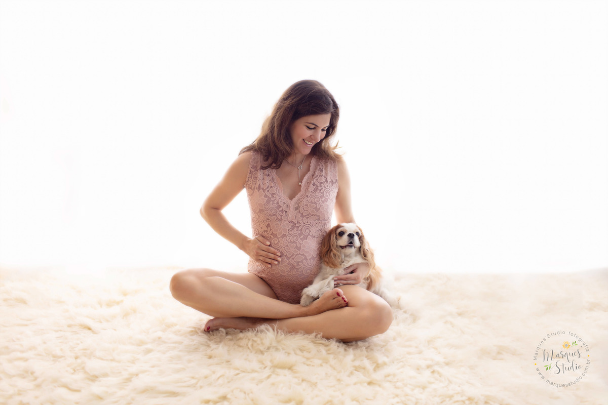 Fotografia tirada em ensaio de gestante feito em studio que encontra-se na Vila Leopoldina - São Paulo, SP. Na foto temos a futura mamãe sentada no chão, com pernas de índio, uma mão na barriga e a outra segurando sua filha canina. 