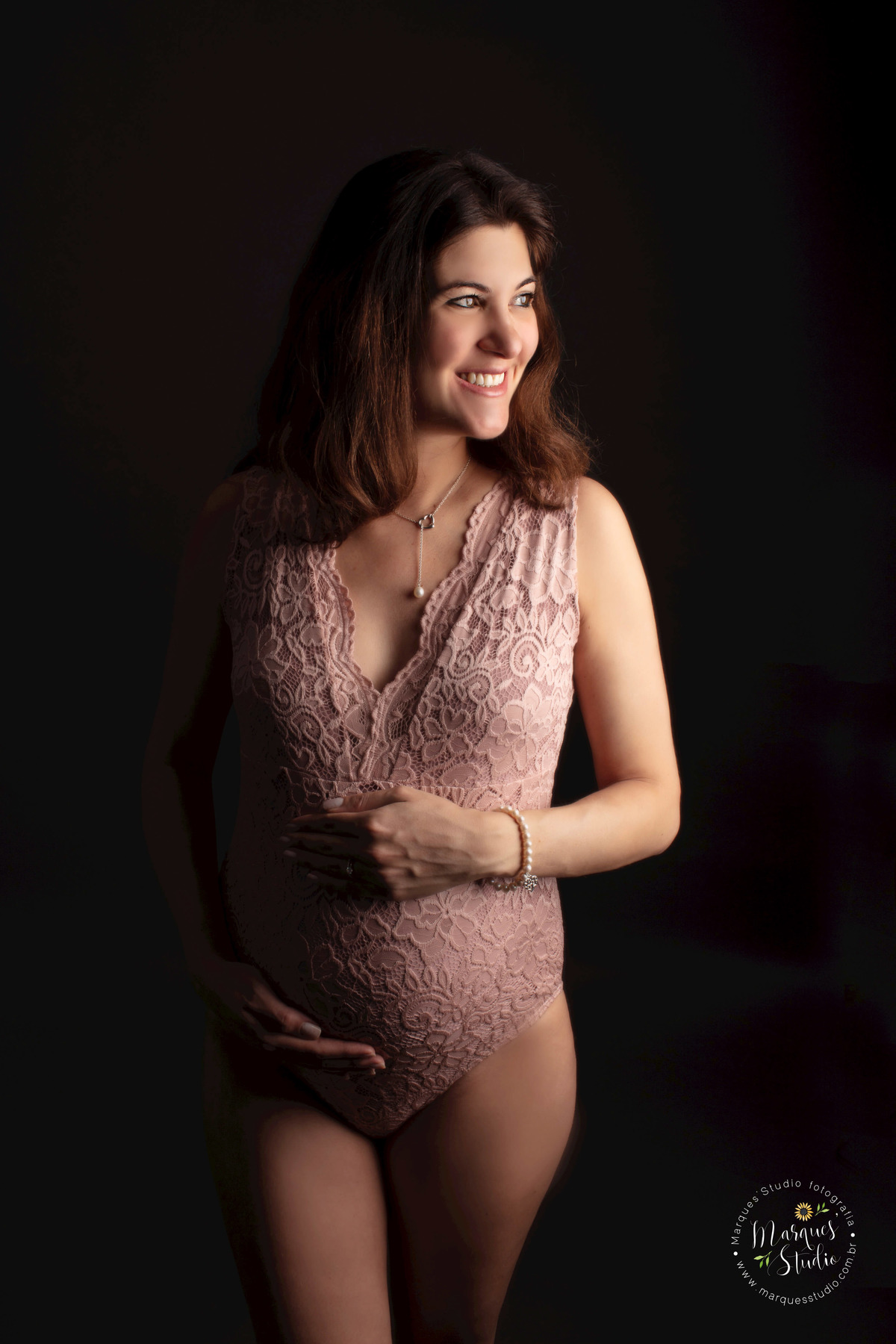 Foto feita no ensaio de gestante da Denise de 32 semanas. Na foto ela está no fundo preto, usando um lindo Bore rosa, sorrindo para a luz enquanto segura sua barriga de gestante. A foto foi feita no studio que encontra-se no Morumbi - São Paulo, SP.