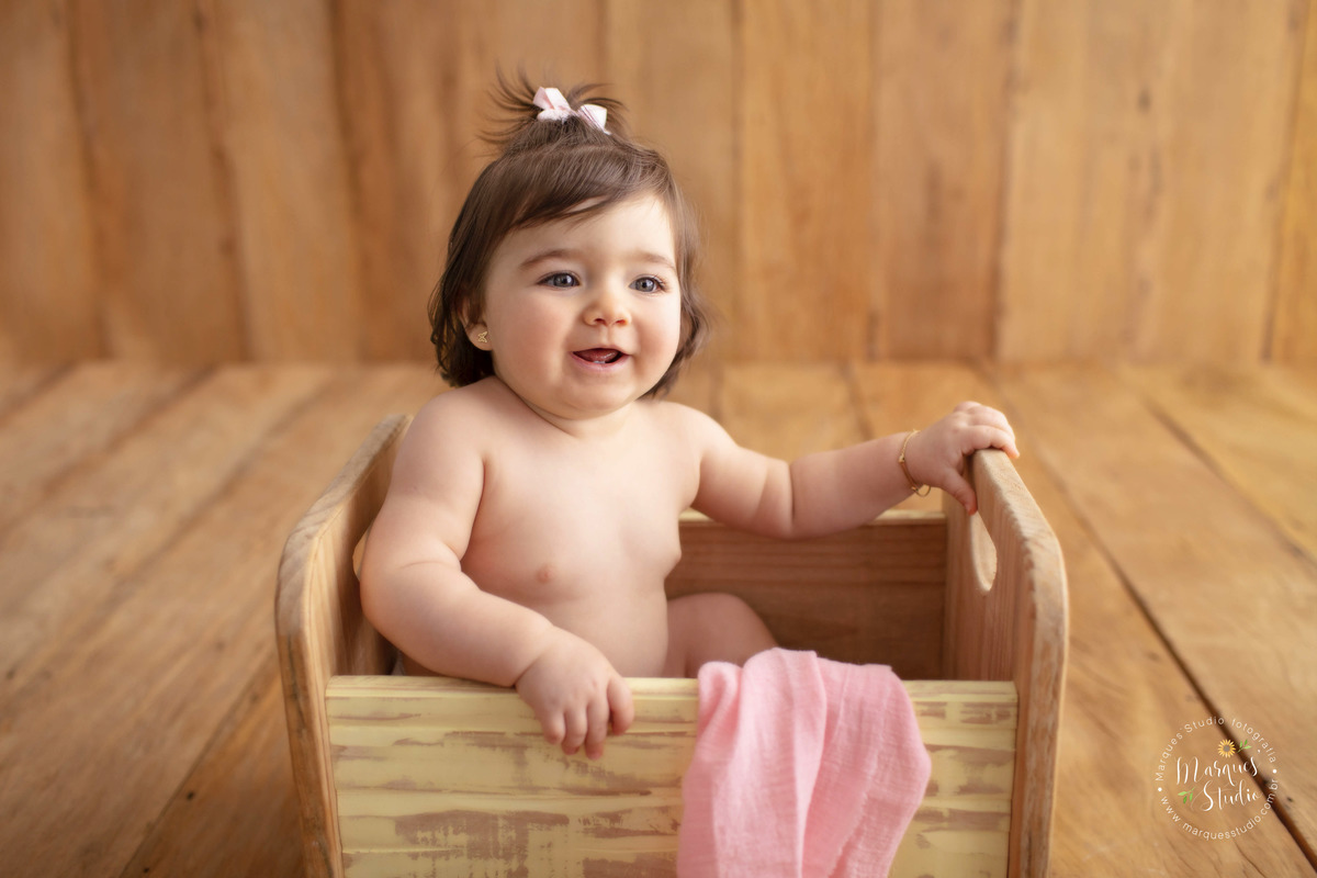 Foto tirada em studio localizado na Zona Sul de São Paulo, SP. Na foto temos a Lorena de 10 meses sentada em um berço de madeira, com um pano rosa ao redor, usando um lindo laço branco na cabeça, ela está muito sorridente, o fundo fotográfico é de madeira