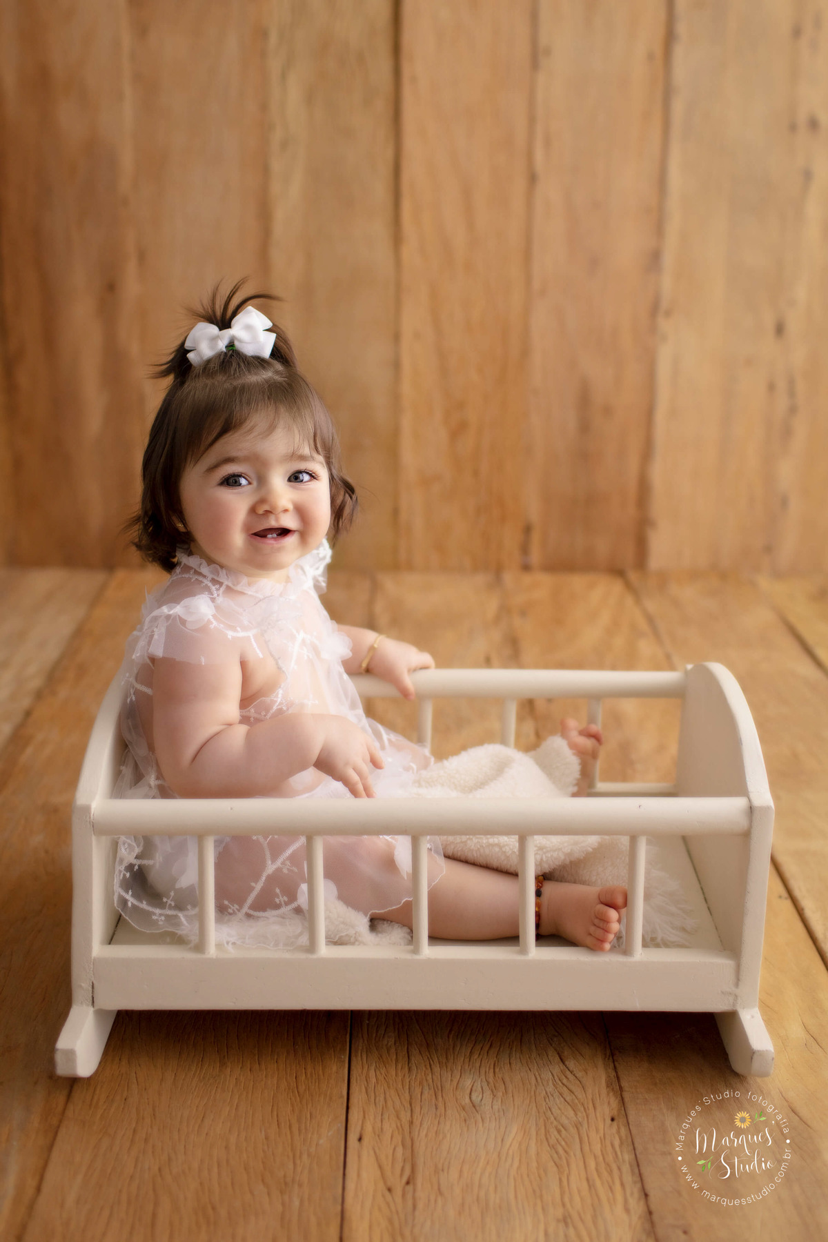 Ensaio fotográfico feito em studio que está localizado na Vila Leopoldina - São Paulo, SP. Na foto temos a Lorena de 10 meses, sentada em um bercinho de madeira branca, usando um lindo vestido e laço também brancos. Ela esta sorridente.