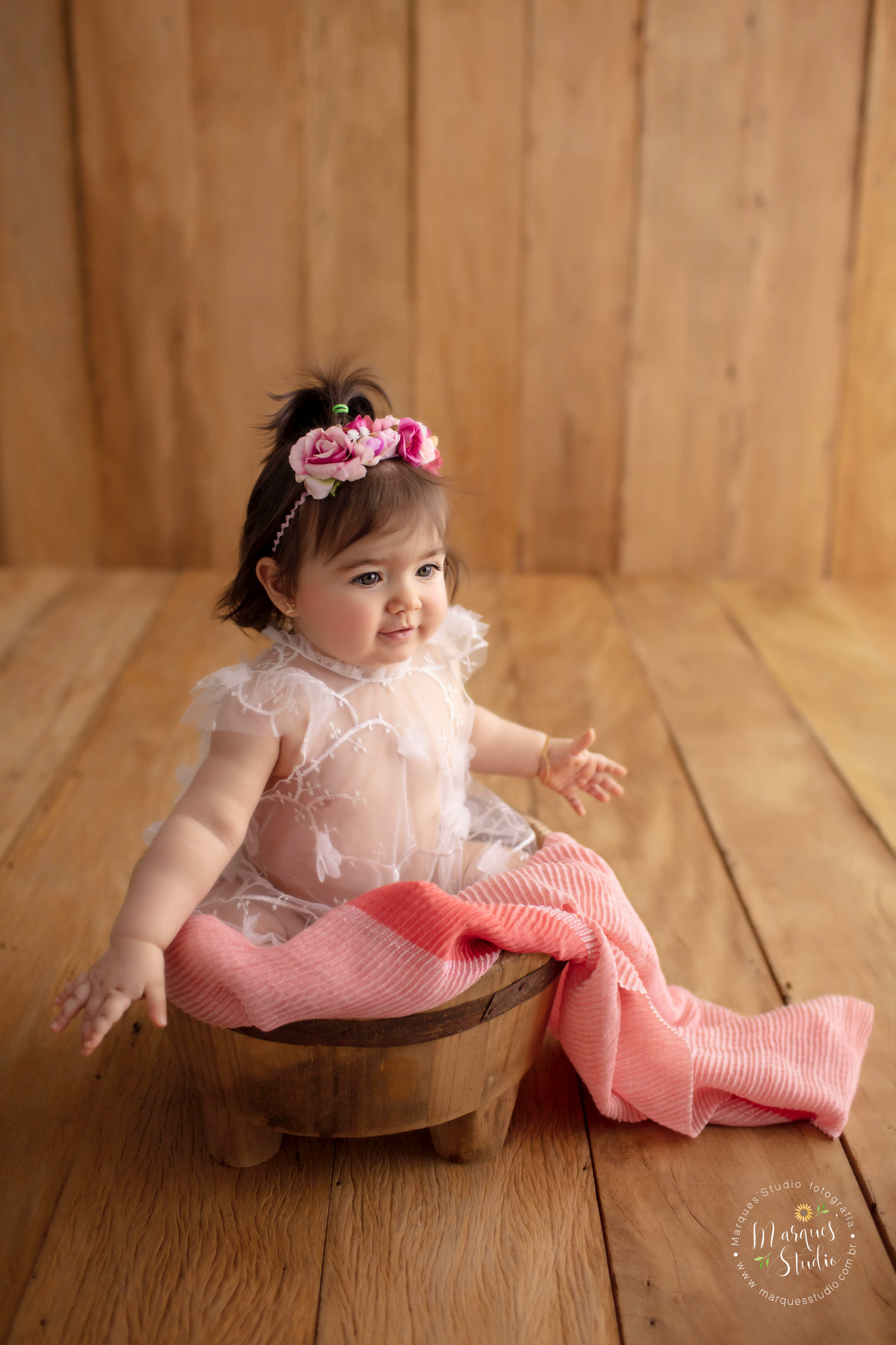 Foto registrada no ensaio da Lorena de 10 meses. Na foto a bebê está sentada em um balde de madeira, com um tecido rosa em volta dele, ela usa um lindo vestido branco e flores na cabeça. O studio encontra-se localizado na Av. Paulista- São Paulo, SP.