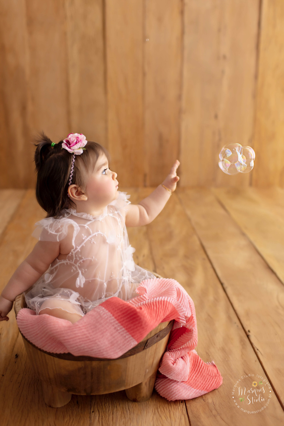 Foto tirada no ensaio da Lorena. Na foto a bebê está sentada em um balde de madeira, com um tecido rosa em volta dele, ela usa um vestido branco e flores na cabeça. Ela está brincando com uma bola de  sabão. O studio está na Av.Consolação - São Paulo, SP.