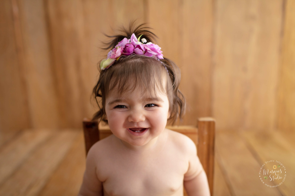 Fotografia feita em ensaio de criança no studio que fica localizado em Osasco - São Paulo, SP. Na foto temos um close do rosto da bebê. Ela está usando um lindo arranjo de flores rosas na cabeça e está dando uma gostosa gargalhada.