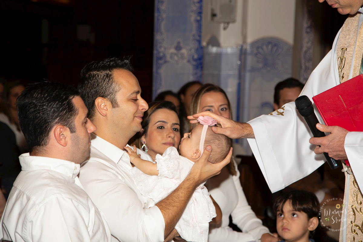 Foto feita no batizado da Martina na Vila Madalena - São Paulo, SP. Na foto a Martina está no colo do seu papai, enquanto recebe as bençãos e orações do Padre, que está com as mãos em sua cabeça.
