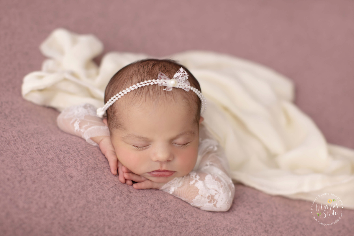 Fotografia registrada em studio localizado no Bela Vista - São Paulo, SP. A bebê está usando uma roupinha branca, com um laço na cabeça e coberta com um tecido branco, deitada em uma manta rosa 