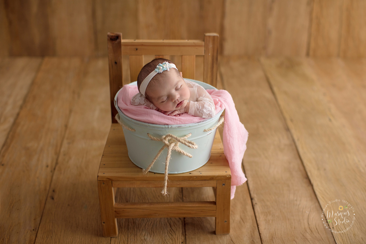 Fotografia registrada em studio localizada no Alto de Pinheiros - São Paulo - SP. Na foto a bebê está em um fundo fotográfico de madeira, deitada de bruços em um cestinho verde em cima de uma cadeira de madeira.