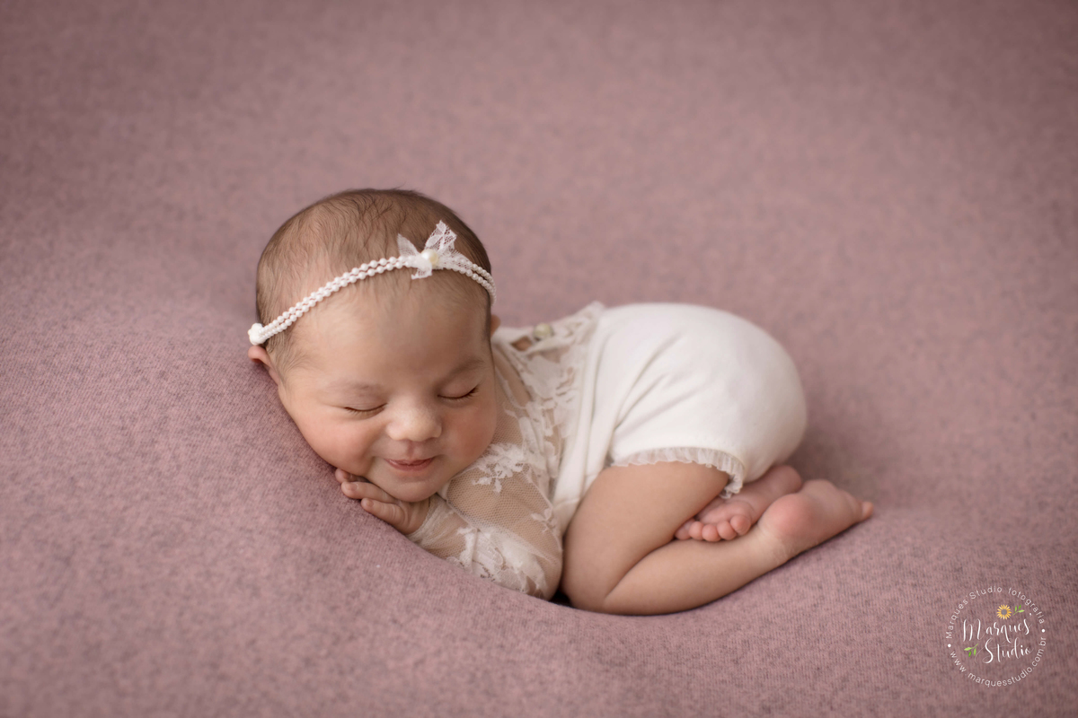 Fotografia registrada em studio localizado no Jardins - São Paulo, SP. A bebê está usando uma roupinha branca, com um laço na cabeça e coberta com um tecido branco, deitada em uma manta rosa 