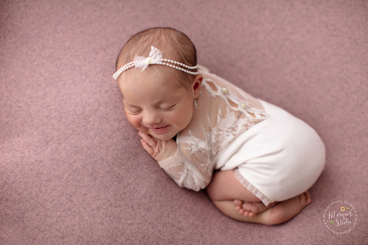Ensaio Newborn da Laura de 11 dias. Na foto a bebê está deitada em um puff rosa, ela usa um macaquinho branco de renda e uma tiara de pérolas, ela está sorrindo. O studio encontra-se localizado na Vila Leopoldina - São Paulo, SP.