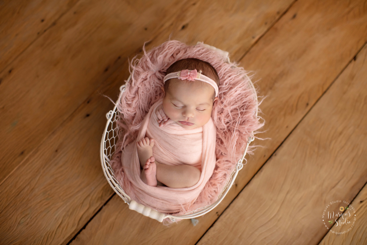 Ensaio newborn da Laura de 11 dias. Na foto a Laura está enrolada em um wrap rosa, em cima de uma cestinha branca com um tecido rosa e o fundo fotográfico é de madeira. O studio encontra-se em Pinheiros - São Paulo, SP.