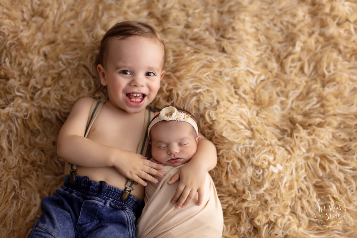 Foto tirada no ensaio newborn da Laura de 11 dias em studio localizado em Perdizes - São Paulo, SP. Na foto a bebê está enrolada em um um wrap salmão enquanto seu irmãozinho mais velho abraça ela, os dois estão deitados em um tapete mesclado.