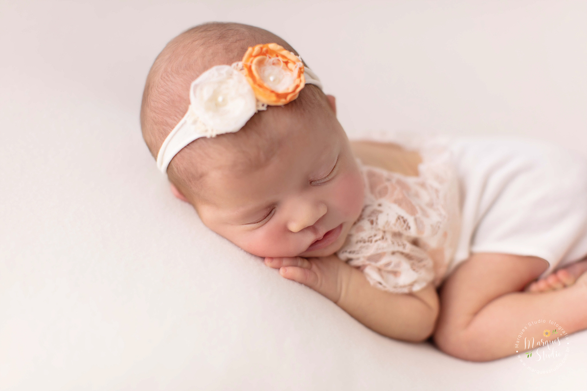 Foto tirada no Newborn da Alice de 18 dias no studio que localiza-se em Osasco- São Paulo, SP. Na foto a bebê está deitada em puff branco, usando um macacão branco de renda e uma tiara de flores na cabeça. Ela está com as mãos em baixo do queixo.