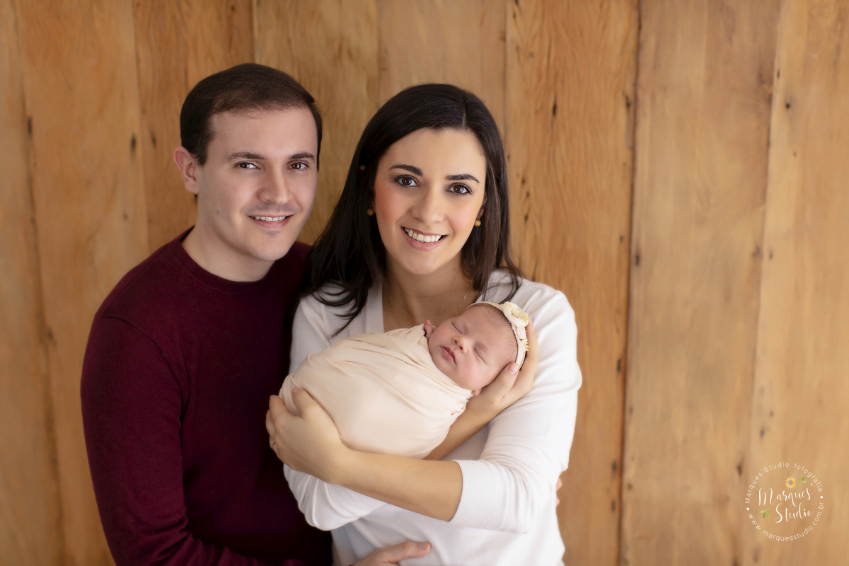 Foto da família da bebê recém nascida Alice em seu ensaio Newborn, realizado em Studio que fica na Zona Sul de São Paulo, SP. Na foto a mamãe segura a Alice no colo enquanto o papai abraça as duas. Todos estão sorrindo
