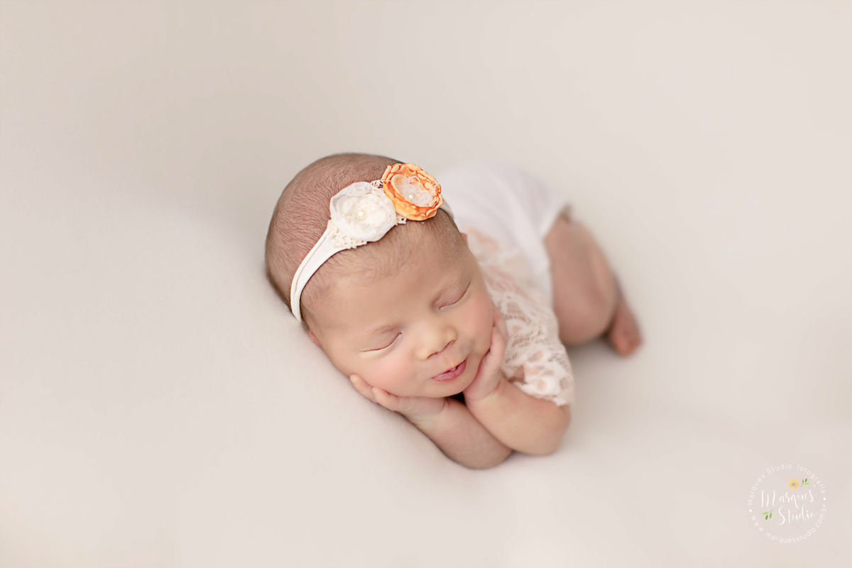 Newborn da Alice de 18 dias em studio localizado na Vila Leopoldina - São Paulo, SP. Na foto a bebê está deitada em puff branco, usando um macacão branco de renda e uma tiara de flores na cabeça. Ela está fazendo biquinho com as mãos em baixo do queixo.