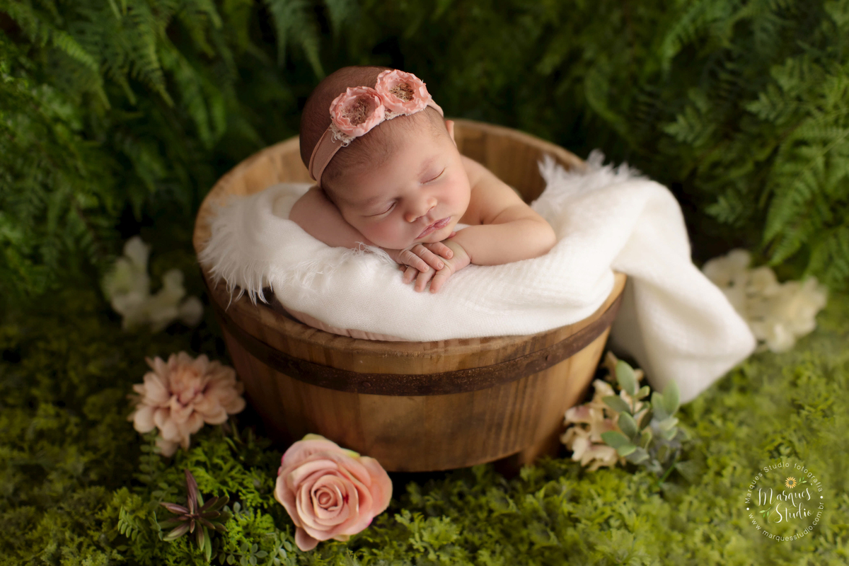 Fotografia registrada em studio que encontra-se no Morumbi - São Paulo, SP. Na foto a bebê Alice está deitada em embalde de madeira que está envolto por um pano branco e ao fundo é um lindo jardim. Ela está com as mãos embaixo do queixo e uma tiara rosa.