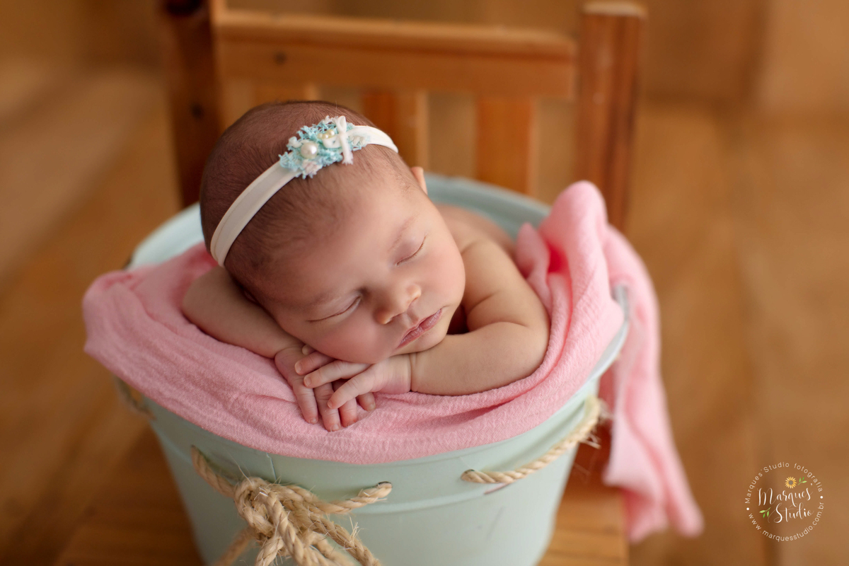 Foto tirada para o ensaio newborn da Alice de 18 dias. Na foto a bebê está em um balde azul, que está envolto por um pano rosa e está em cima de uma cadeira de madeira. Ela está com as mãos em baixo do queixo. O studio encontra-se em Perdizes - São Paulo