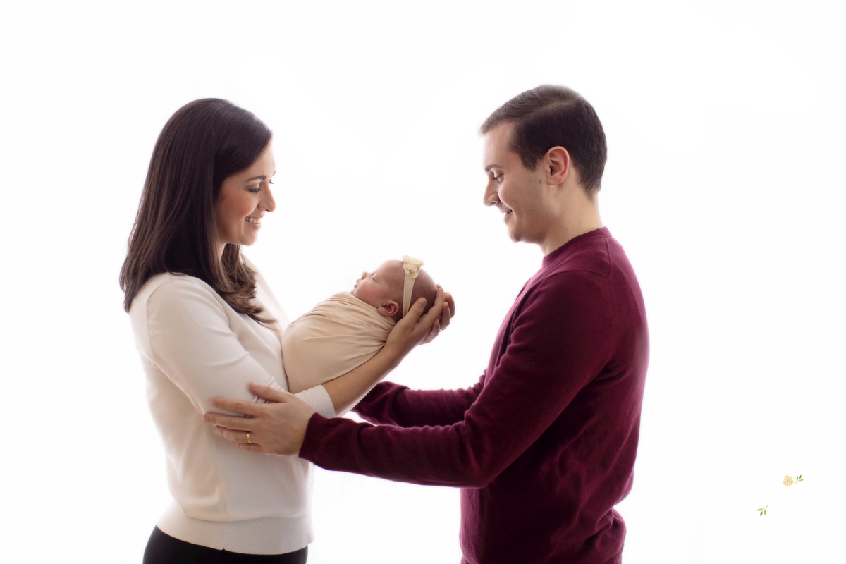 Foto da família da bebê recém nascida Alice em seu ensaio Newborn, realizado em Studio que fica na Zona Oeste de São Paulo, SP. Na foto a mamãe segura a Alice no colo enquanto o papai sorri de frente para as duas.