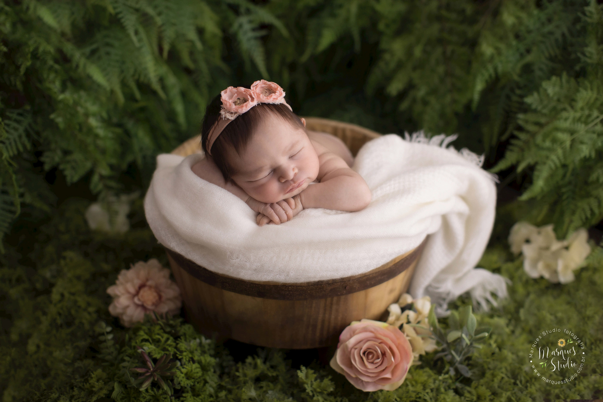 Ensaio newborn da Manuela de 7 dias. na foto a bebê está deitada em um tronco de madeira, usando uma tiara de flores rosa e o fundo fotográfico é um lindo jardim florido. O studio encontra-se na Zona Oeste de São Paulo, SP.