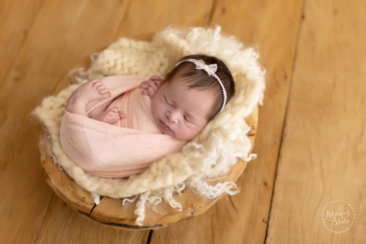 Fotografia tirada em studio que localiza-se no Itaim Bibi- São Paulo, SP. Na foto a bebê está enrolado em um wrep rosa, deitada em um tronco de madeira e usando uma tiara de laço branco. O fundo fotográfico é de madeira. 