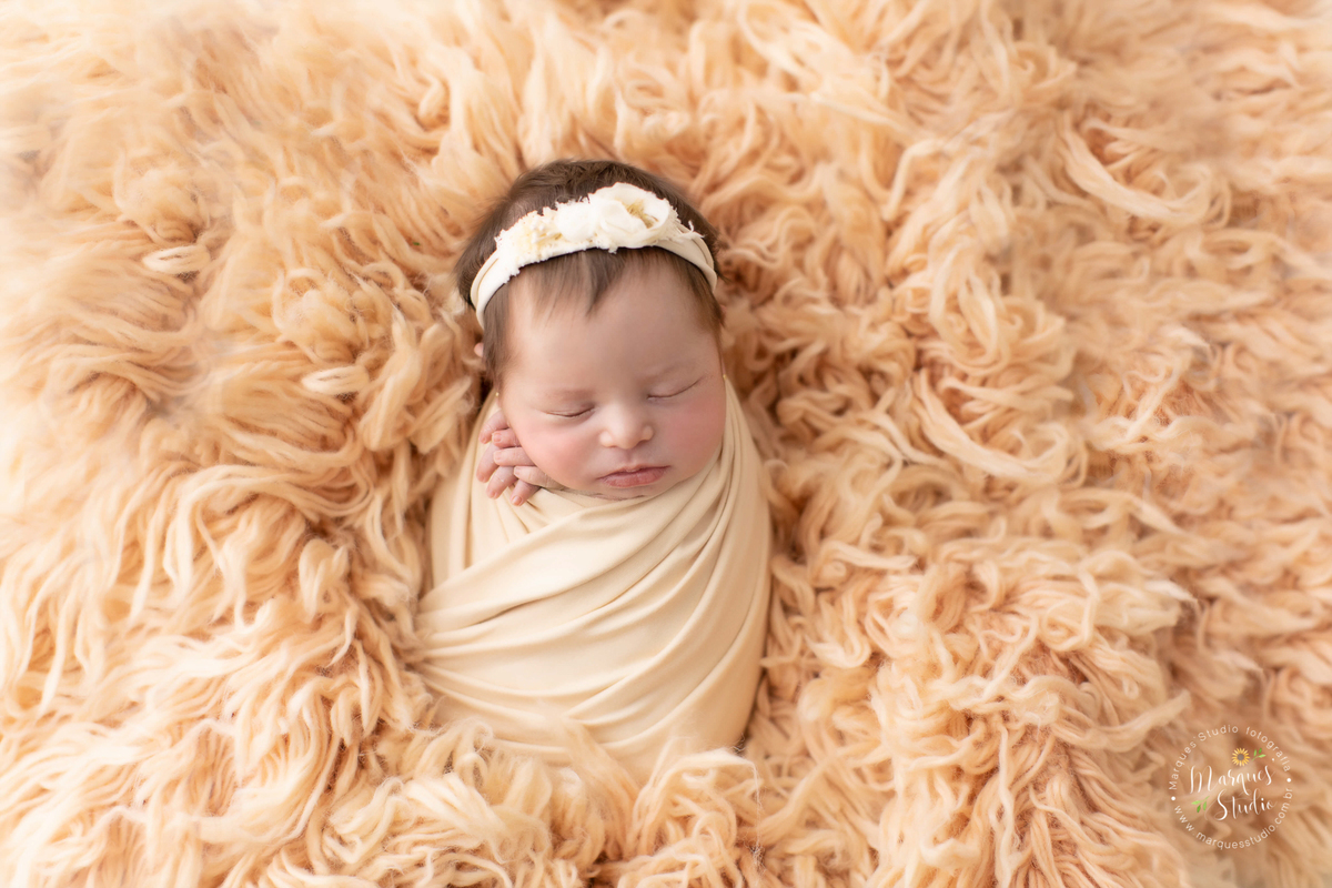 Fotografia tirada em studio que localiza-se na Faria Lima - São Paulo, SP. Na foto a bebê está enrolado em um wrep Salmão, deitada em um tapete salmão e usando uma tiara flores bege.